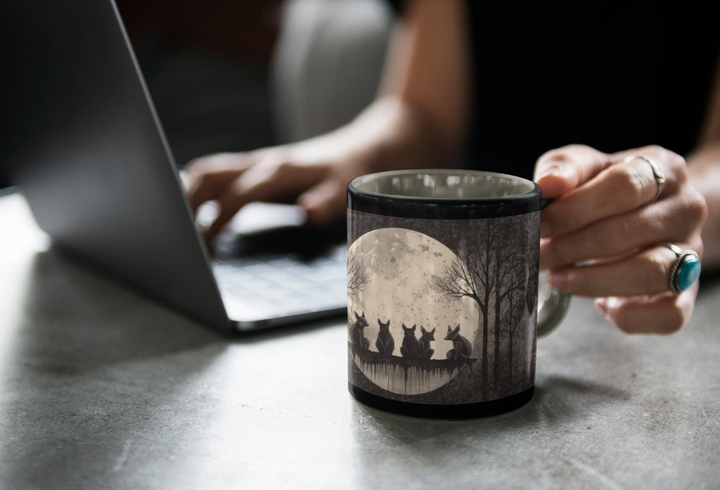 a woman sitting at a desk holding a black coffee mug with an illustration of foxes sitting in front of a full moon in a forest printed on it.