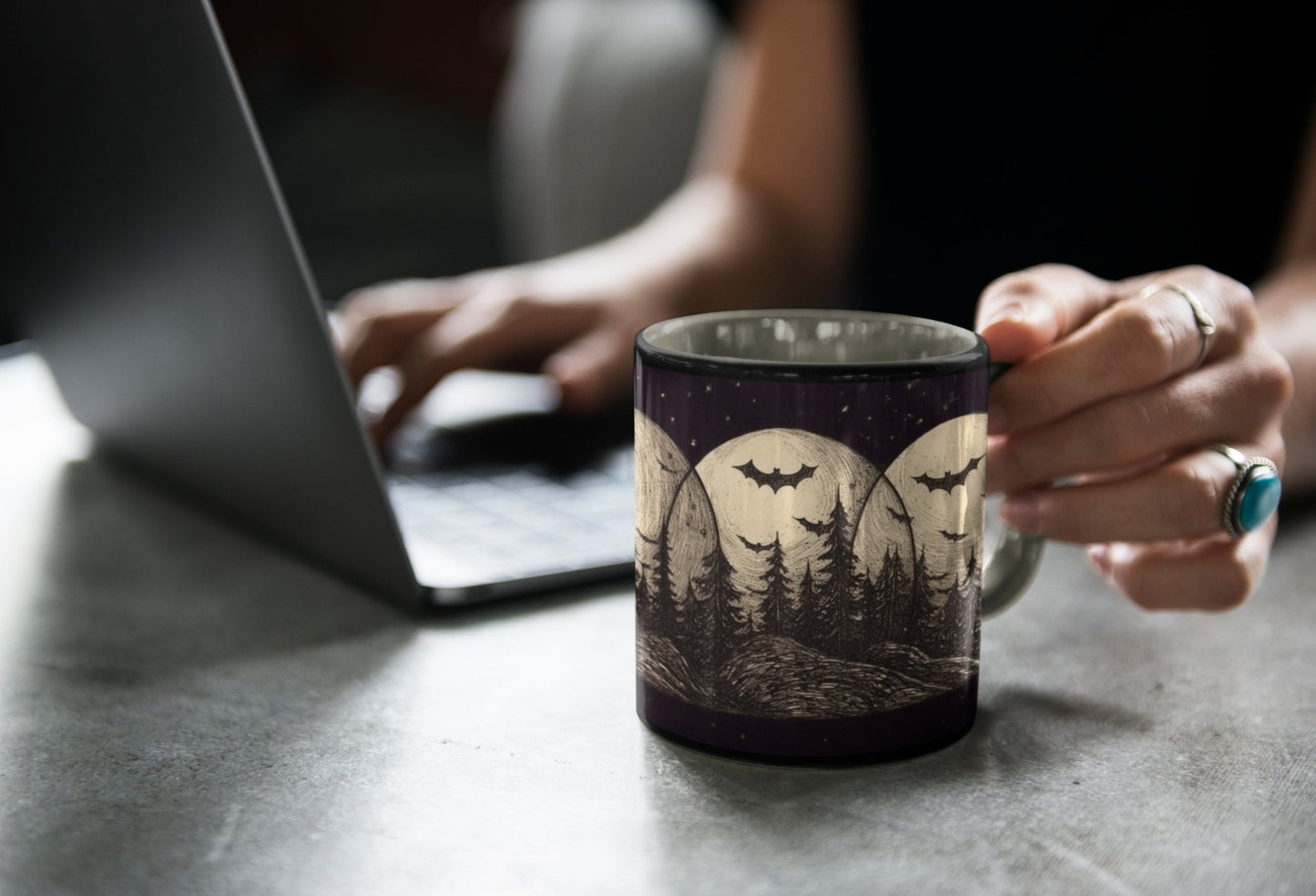 A woman sitting at a desk holding an alternative fashion black coffee mug with an image of bats flying in a forest at night in front of three moons.